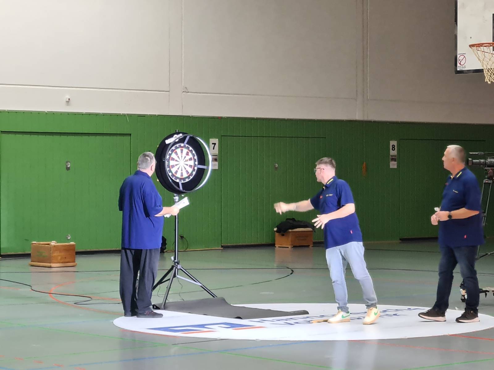 Auf dem Bild sind drei Männer zu sehen, die in einer Turnhalle Dart spielen.