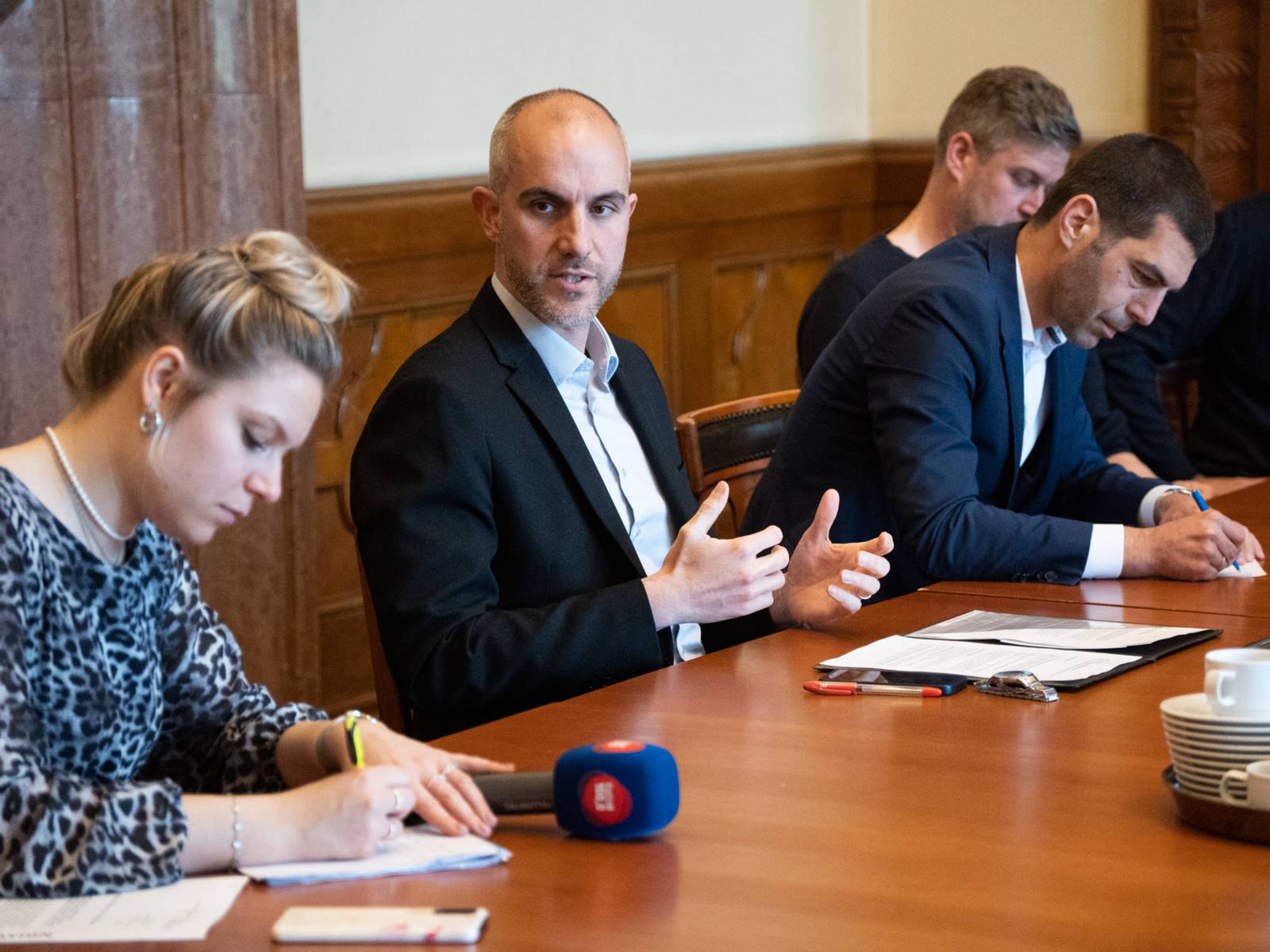 Oberbürgermeister Belit Onay präsentiert das Programm für die bahnhofsnahen Plätze. Er sitzt an einem großen Tisch mit Vertretern der Stadt, weiteren Beteiligten und Journalist*innen.