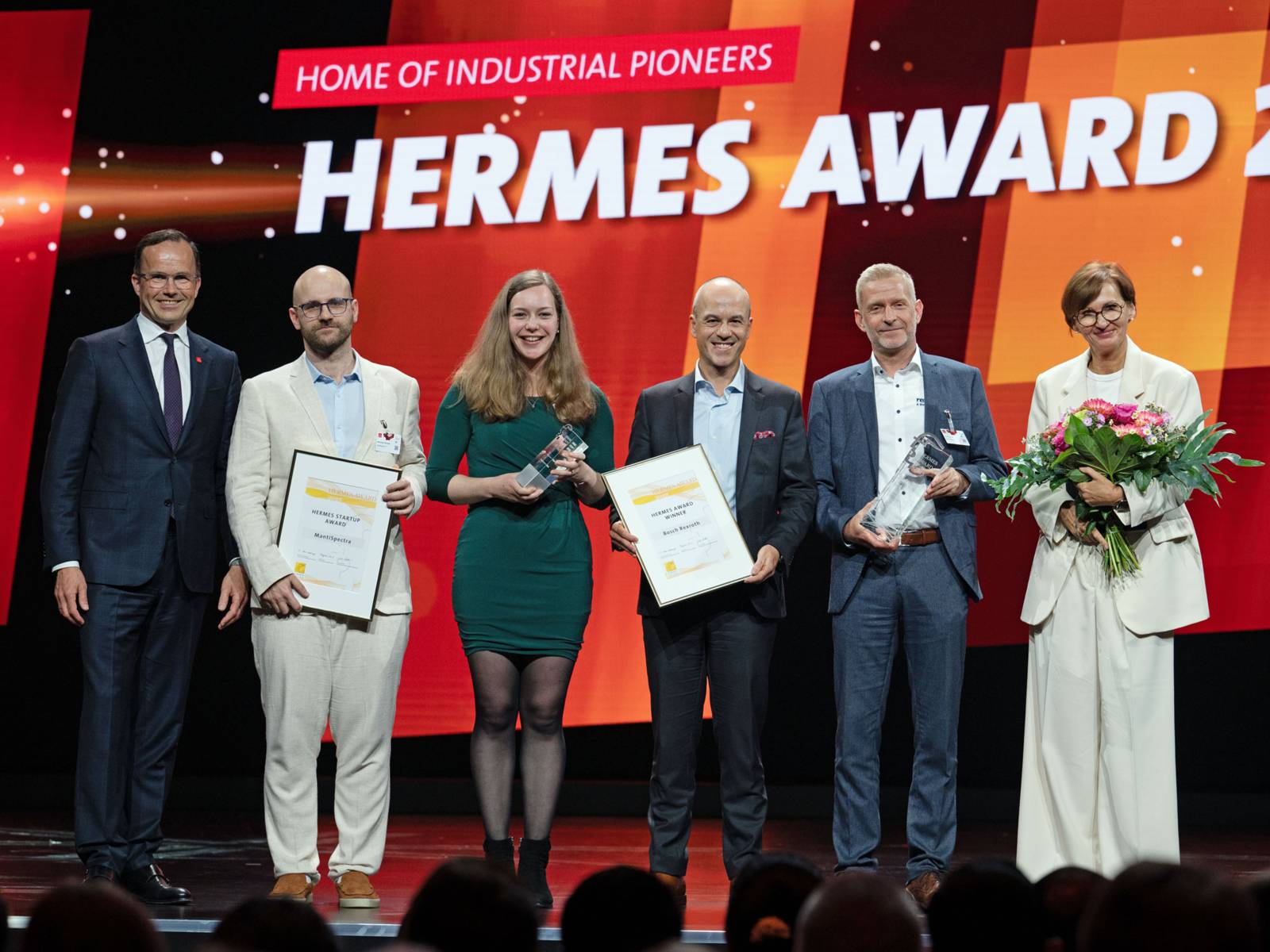 Zwei Frauen und vier Männer posieren mit Urkunden, Pokalen und Blumensträußen auf einem Podium.