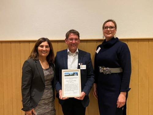 Auf dem Bild sind zwei Frauen (rechts und links) und ein Herr (mitte) zu sehen. Der Herr trägt etwas wie eine Urkunde in der Hand.